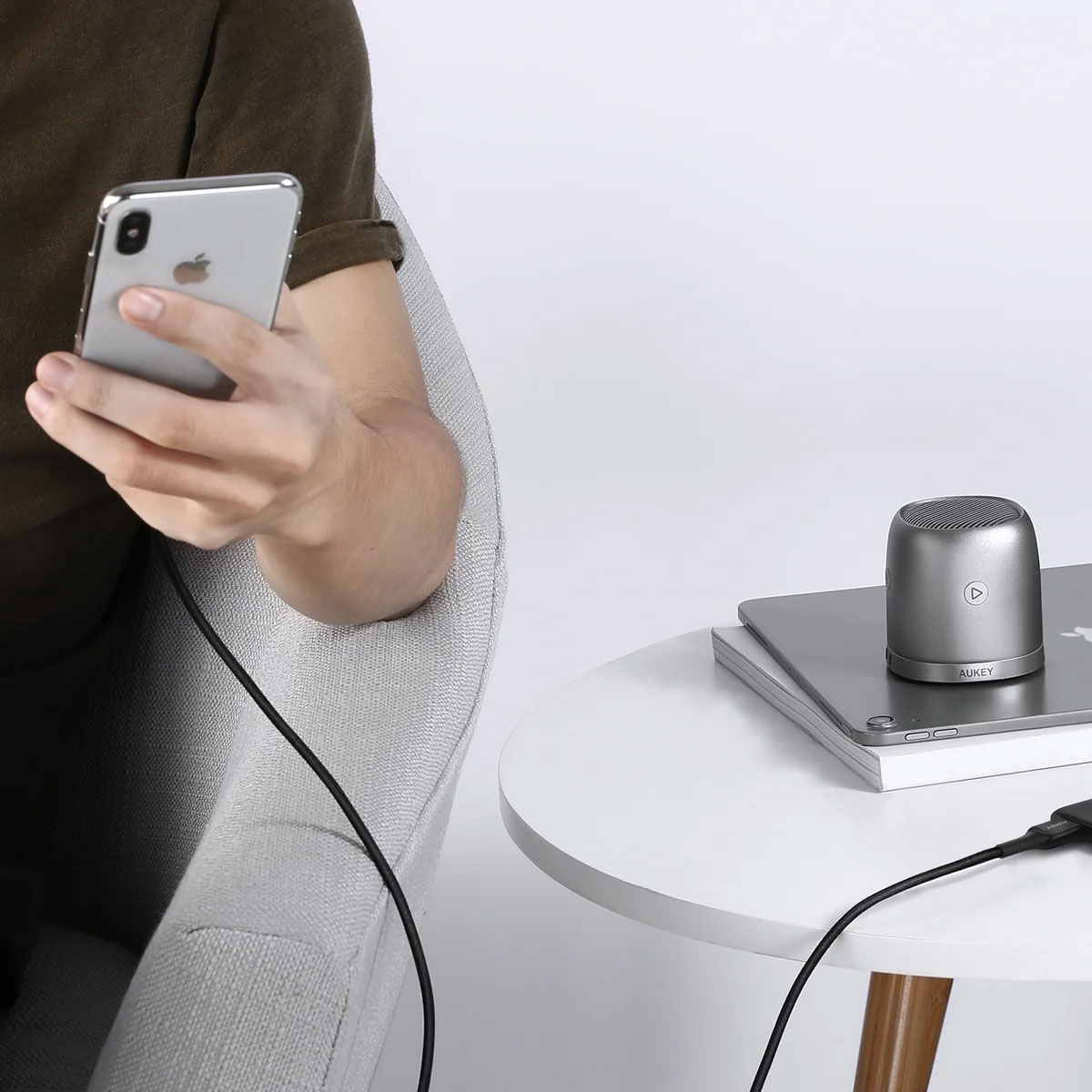Bir sandalyede oturan kişi akıllı telefon kullanıyor. Beyaz masanın üzerinde kablolu küçük gümüş renkli bir hoparlör var.