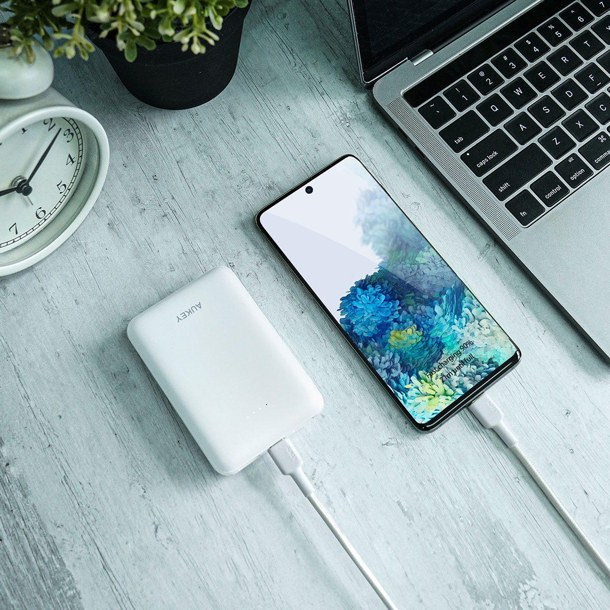 Ahşap masada dizüstü bilgisayar, saat ve bitkinin yanında powerbankten şarj olan beyaz bir akıllı telefon.