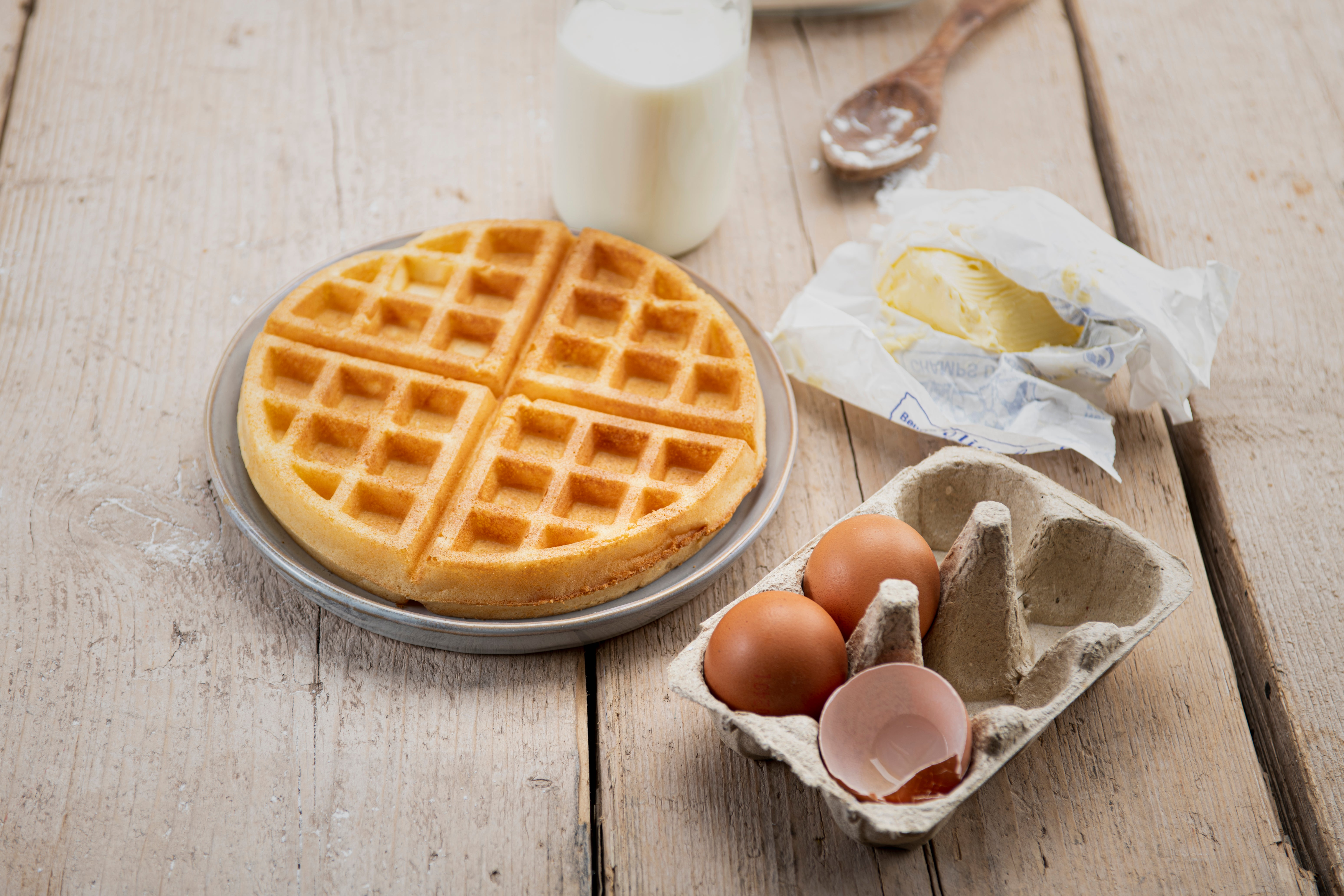 Fresh waffle on a plate with milk, butter, eggs, and a wooden spoon on a wooden table.