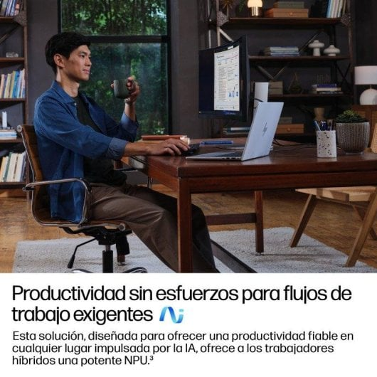 Un hombre está sentado en un escritorio de madera, bebe de una taza y trabaja frente a un portátil y monitor.