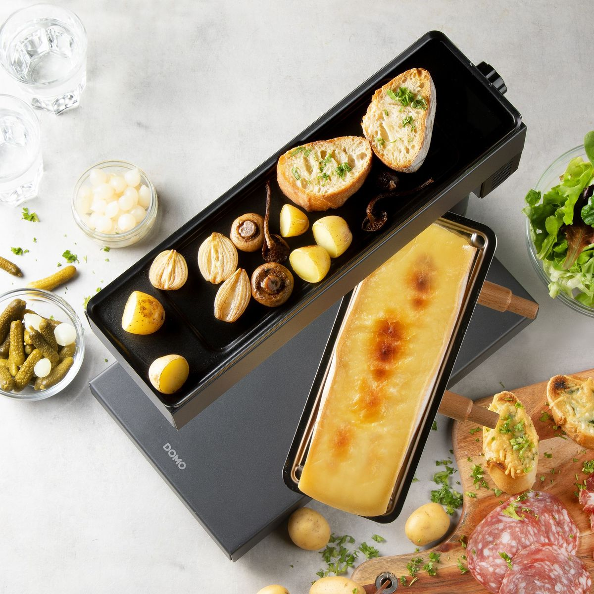 Ein Raclette-Grill mit geschmolzenem Käse, Kartoffeln, Pilzen und Brot, umgeben von Gurken und Salat.