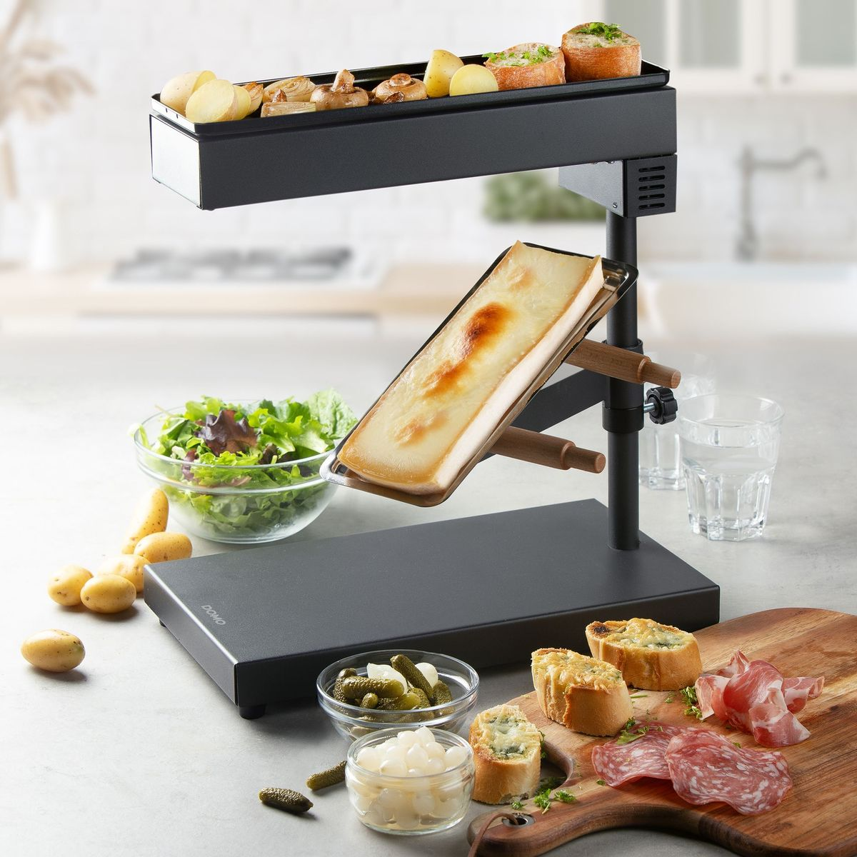 Raclette-Gerät auf Tisch mit geschmolzenem Käse, Kartoffeln, Brot, Wurst und Salat in heller Küche.