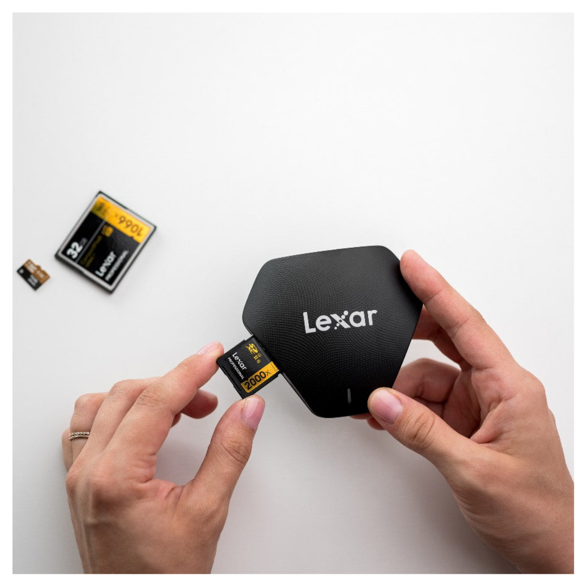 Person steckt eine Lexar-SD-Karte in ein schwarzes Lexar-Lesegerät auf weißem Tisch.