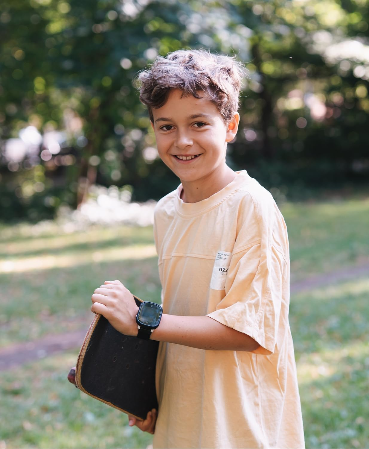 Un niño sonriente con un monopatín en un parque, vistiendo camiseta amarilla y reloj.
