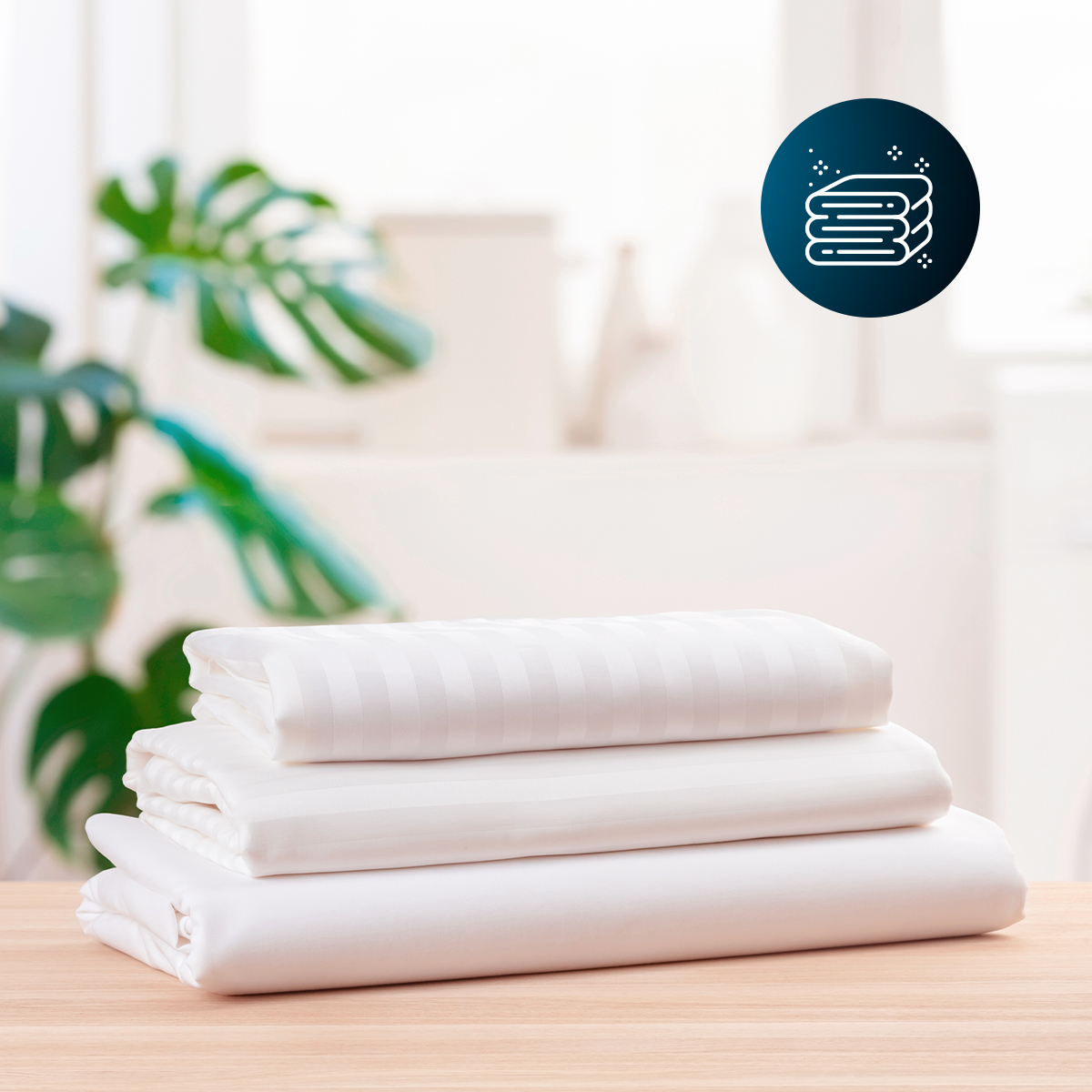 Una pila de ropa de cama blanca sobre una mesa de madera, con una planta de interior al fondo.