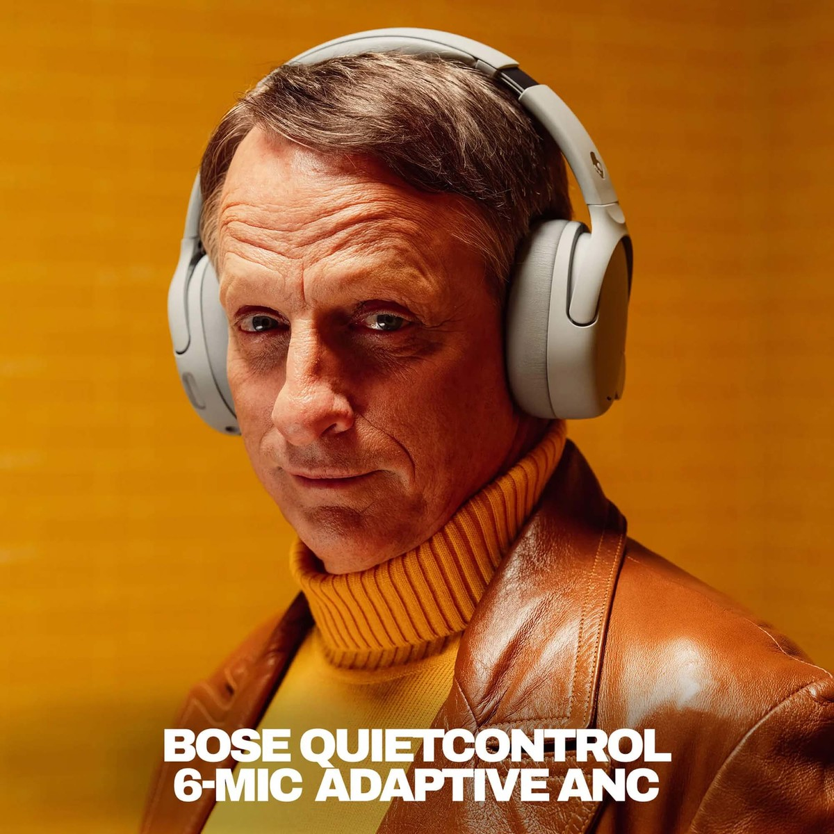 Hombre con auriculares grises, chaqueta de cuero marrón y cuello alto amarillo sobre fondo amarillo.