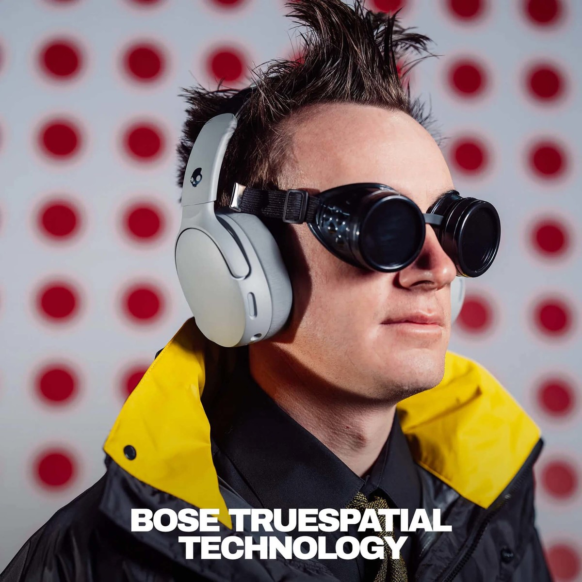 Un hombre con el pelo de punta, gafas y auriculares ante una pared de lunares rojos y blancos.