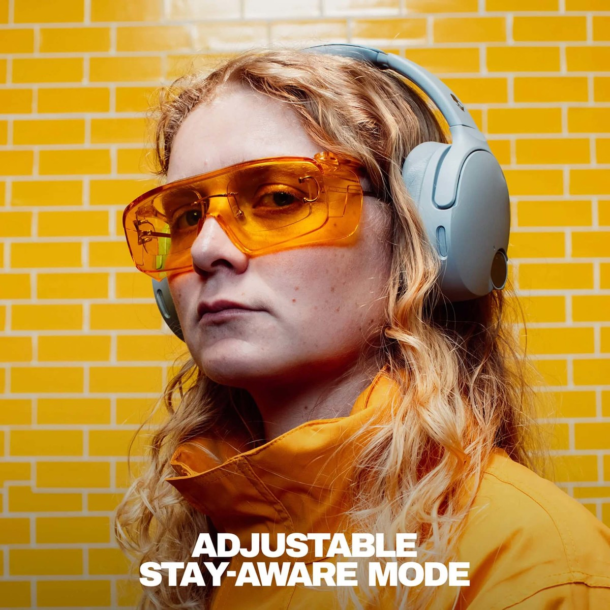 Una mujer rubia con gafas de seguridad naranjas y auriculares grises frente a una pared de ladrillo amarillo.