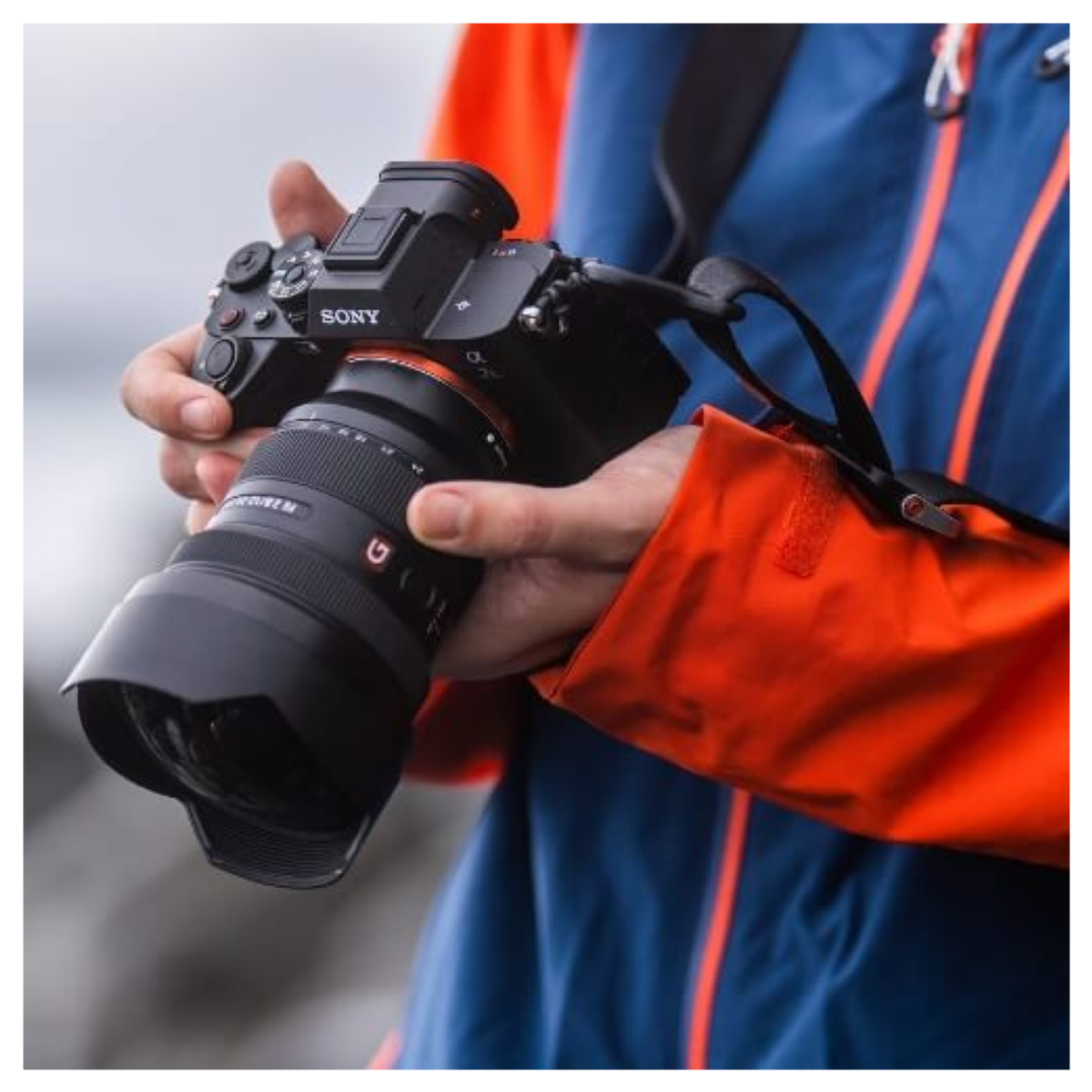 Una persona con una giacca blu e arancione tiene in mano una fotocamera Sony nera con un grande obiettivo zoom.