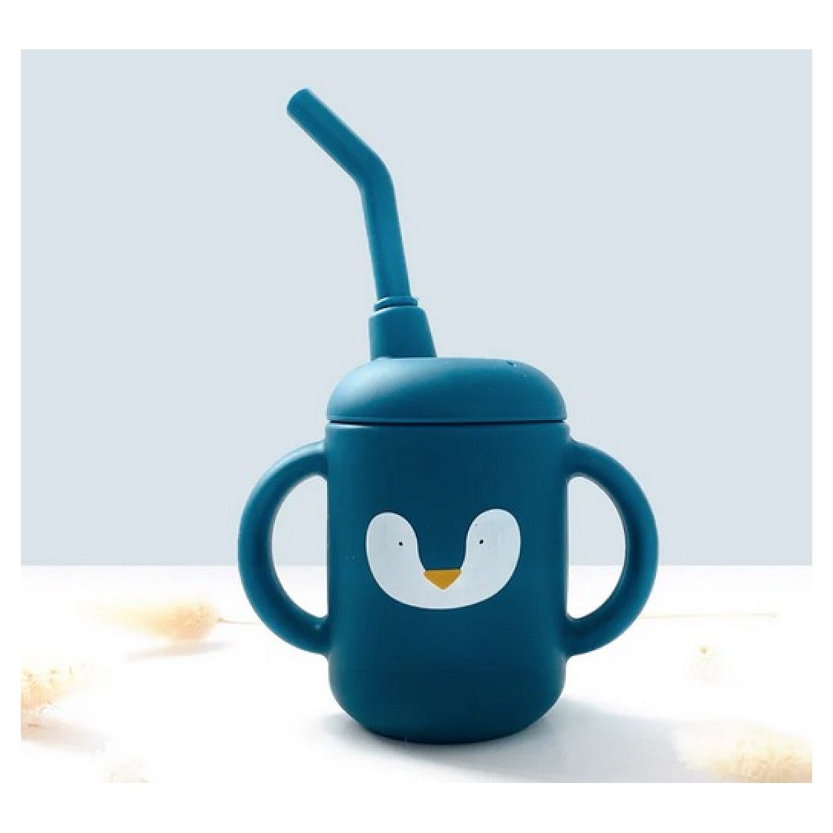 Ein blauer Kinderbecher mit zwei Griffen, Deckel und Strohhalm, verziert mit einem weißen Pinguingesicht.