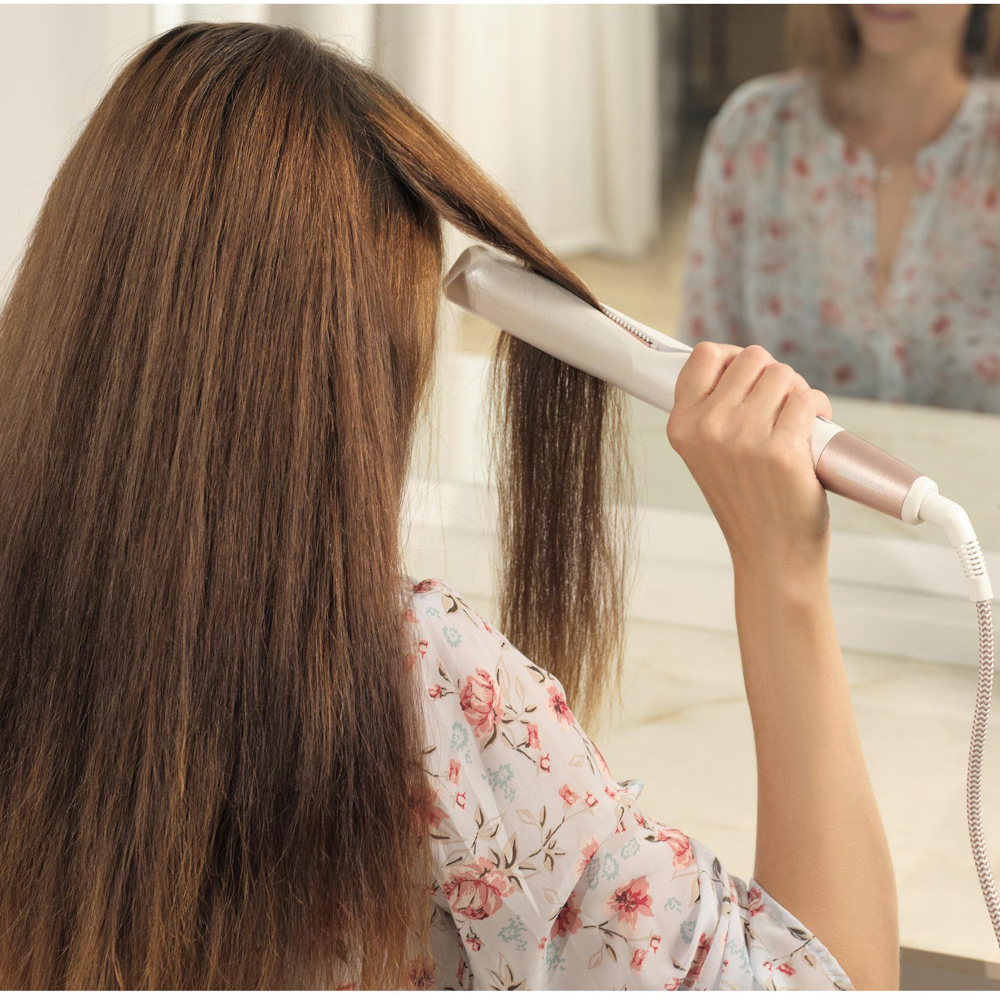 Una donna liscia i suoi capelli castani davanti a uno specchio con una piastra bianca.