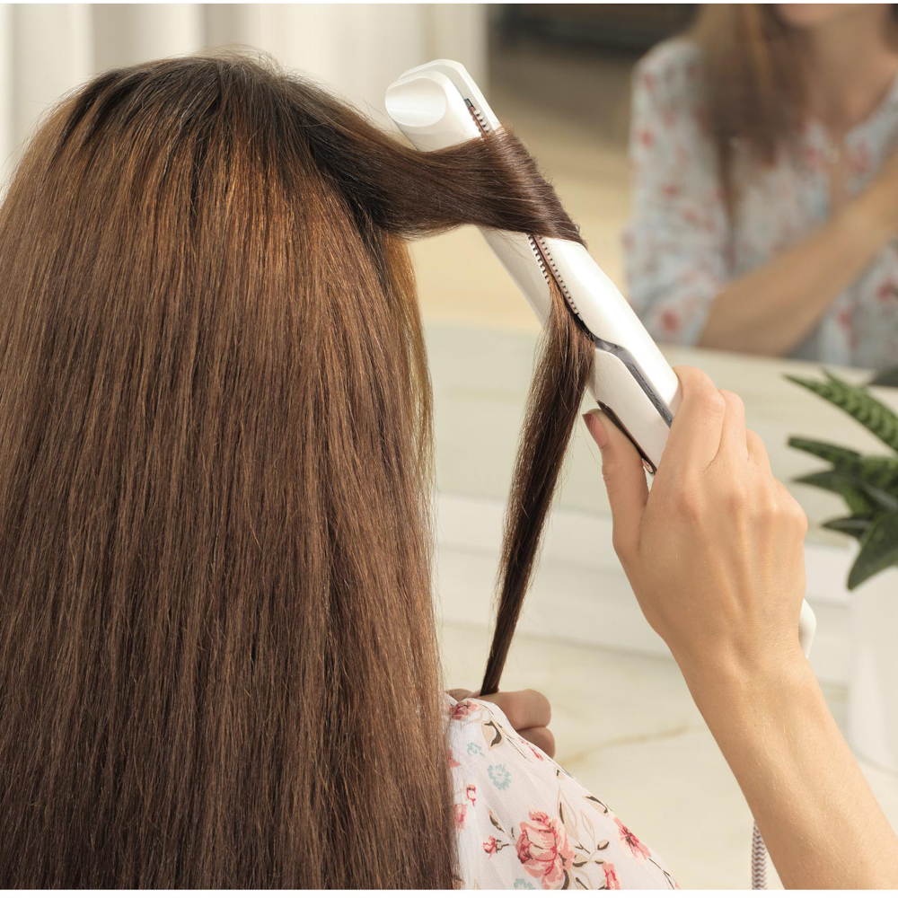 Una donna si stira i capelli castani con una piastra bianca davanti a uno specchio.