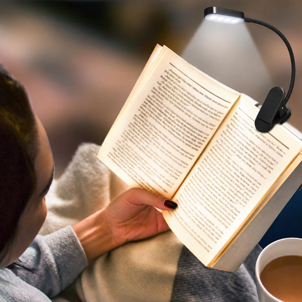 Person reading a book in bed, lit by a small black clip-on lamp. Mug of tea in the foreground.