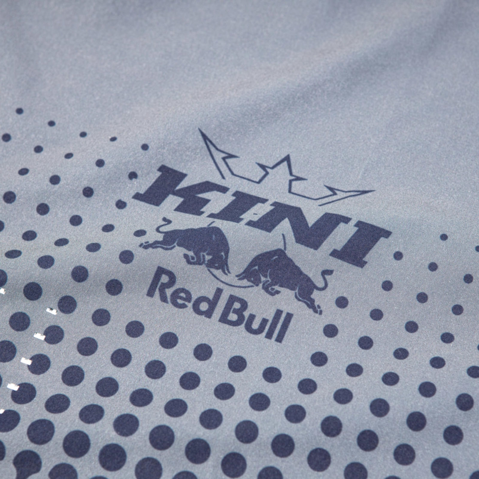 Nahaufnahme eines grauen Stoffes mit dem dunkelblauen Kini Red Bull Logo und Punktverlauf.