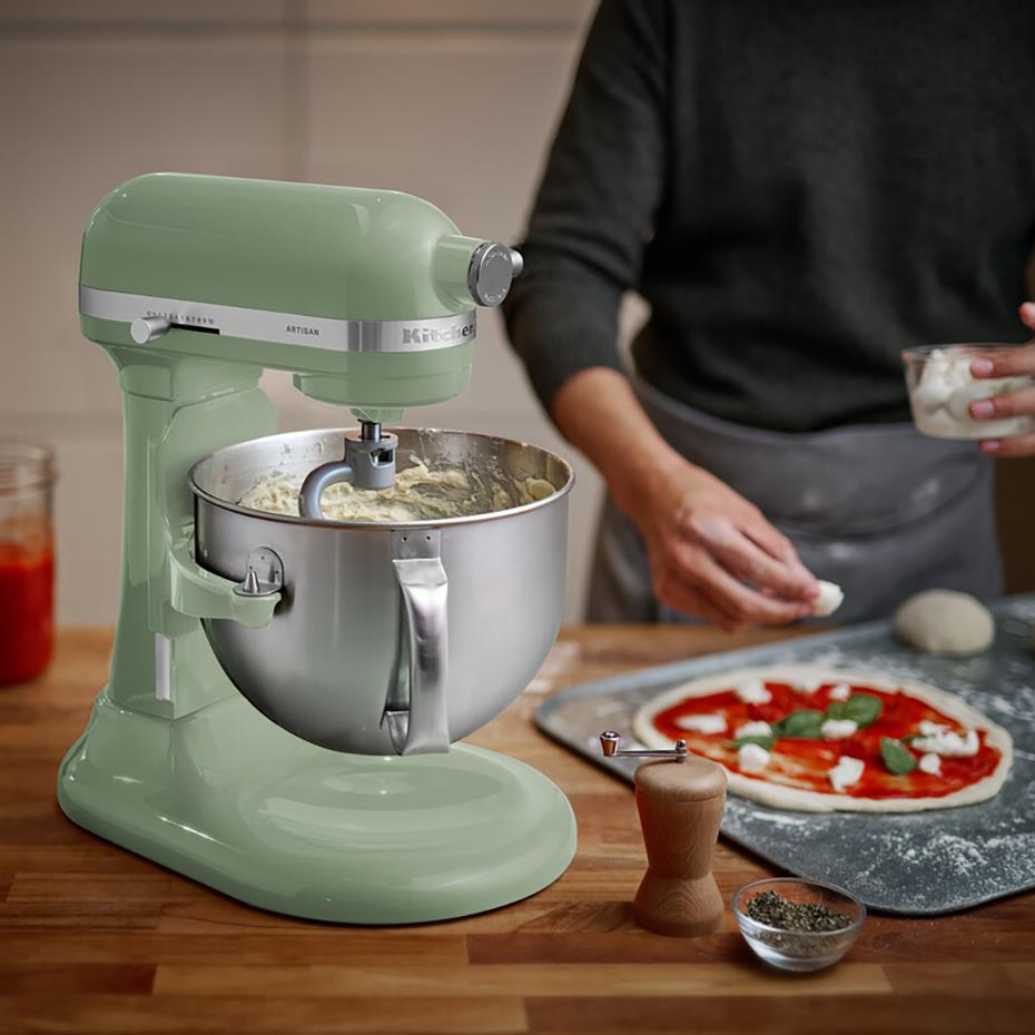 Ahşap tezgah üzerinde açık yeşil stand mikser ve arka planda pizza hazırlayan bir kişi.