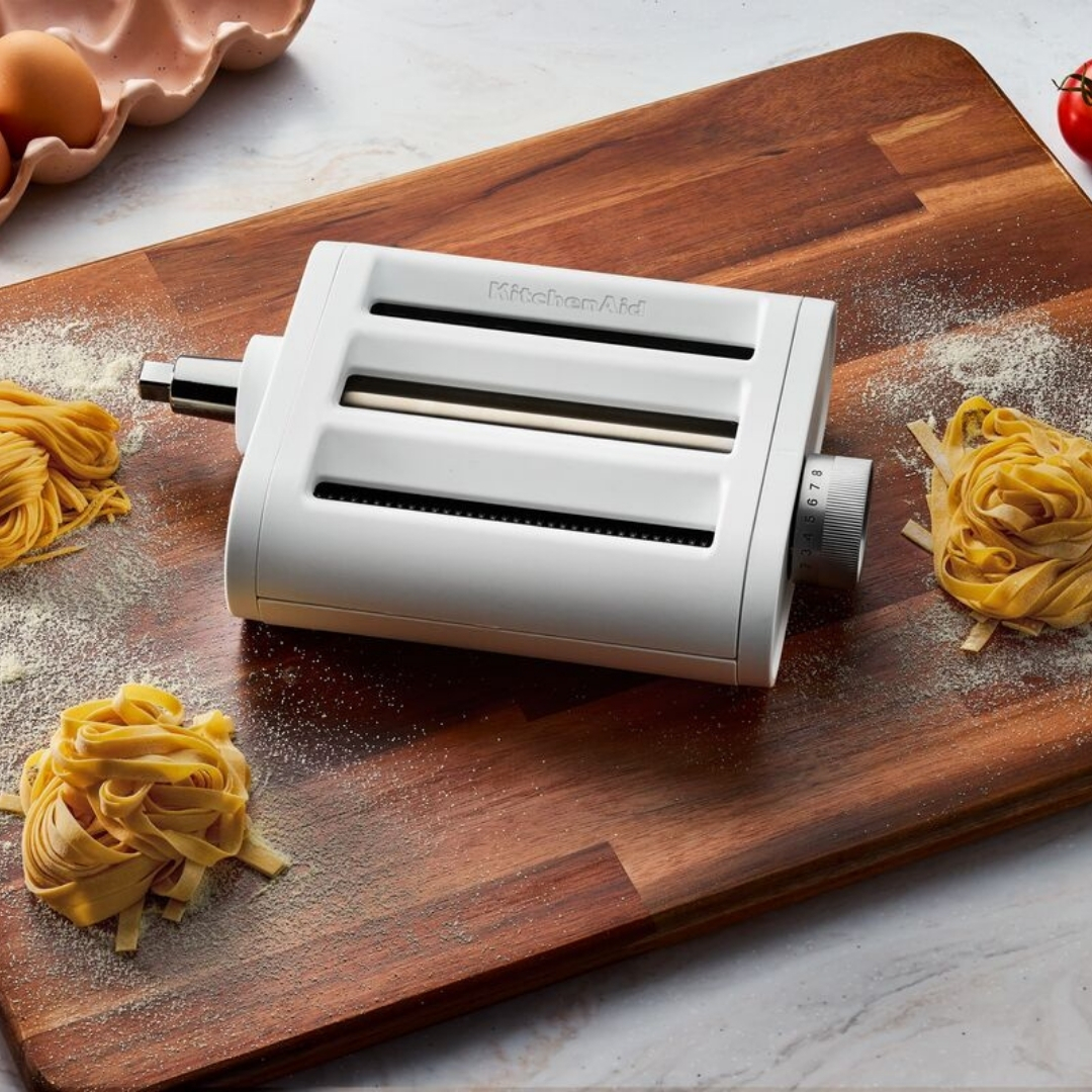 Tahta bir kesme tahtası üzerinde, çiğ makarna, yumurta ve unla çevrili beyaz bir KitchenAid makarna aparatı.