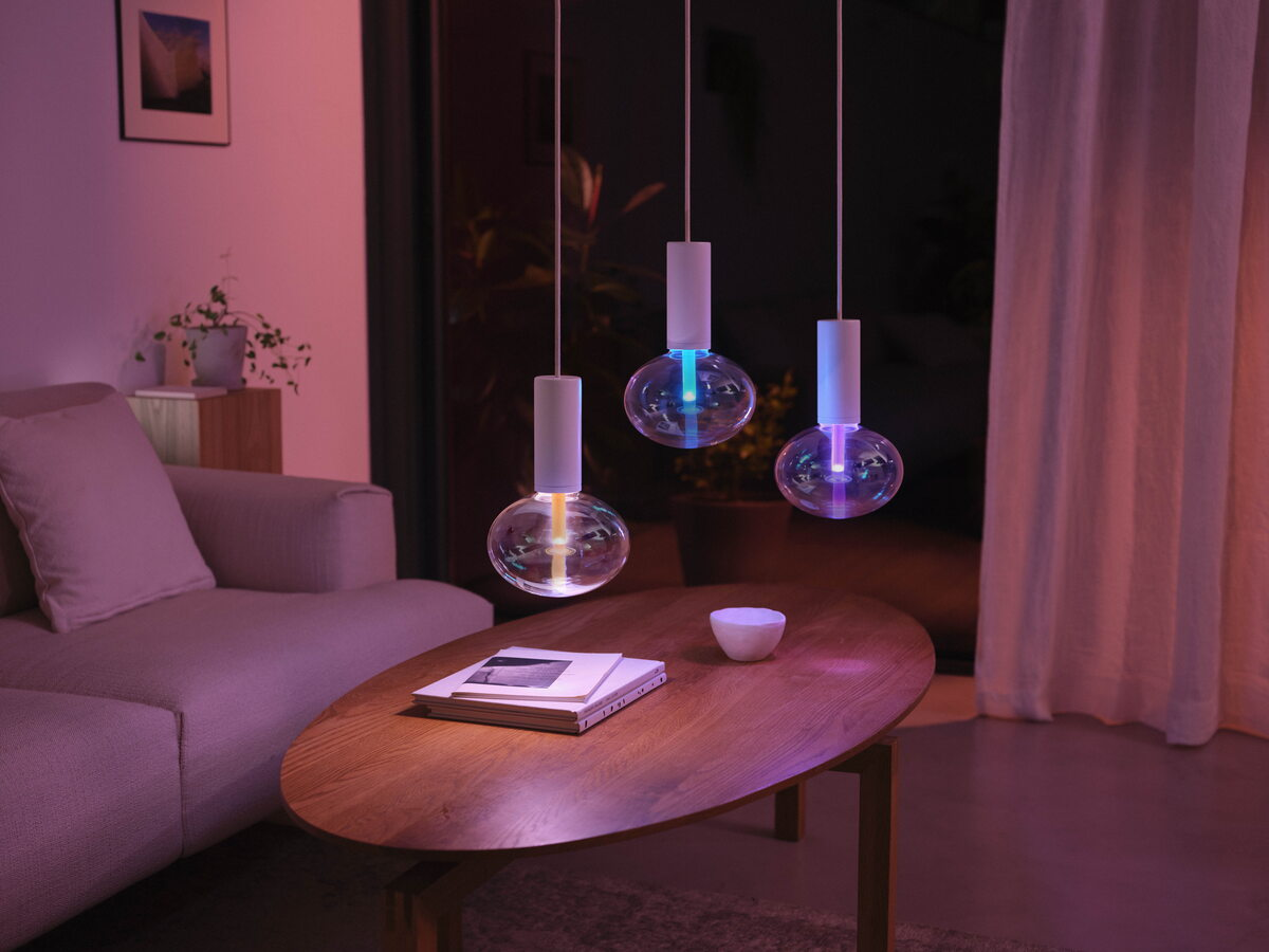 Drei moderne, farbig leuchtende Pendellampen hängen über einem Holztisch in einem gemütlichen Wohnzimmer.