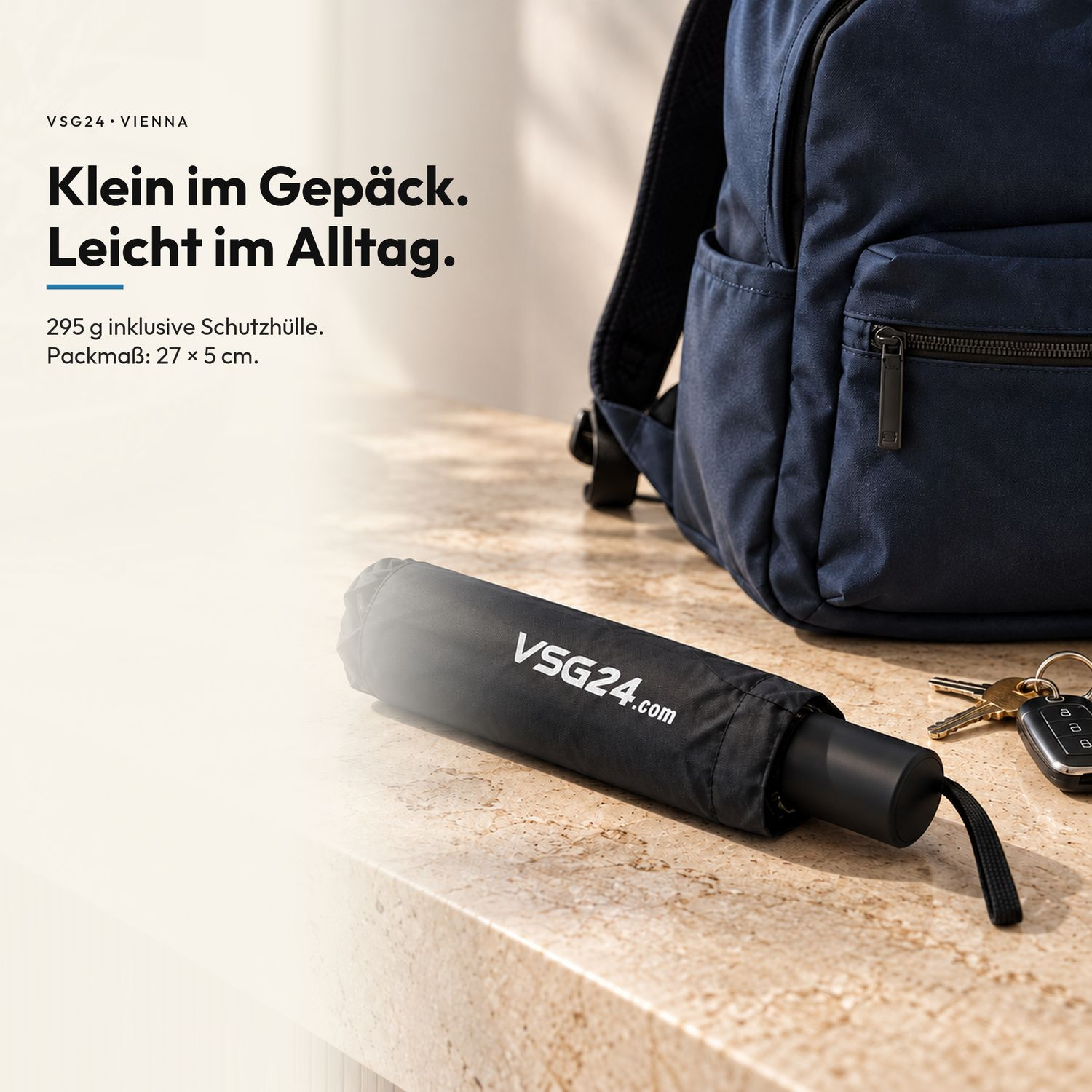 Ein kompakter schwarzer Regenschirm mit Aufschrift liegt auf einer hellen Oberfläche neben einem blauen Rucksack.