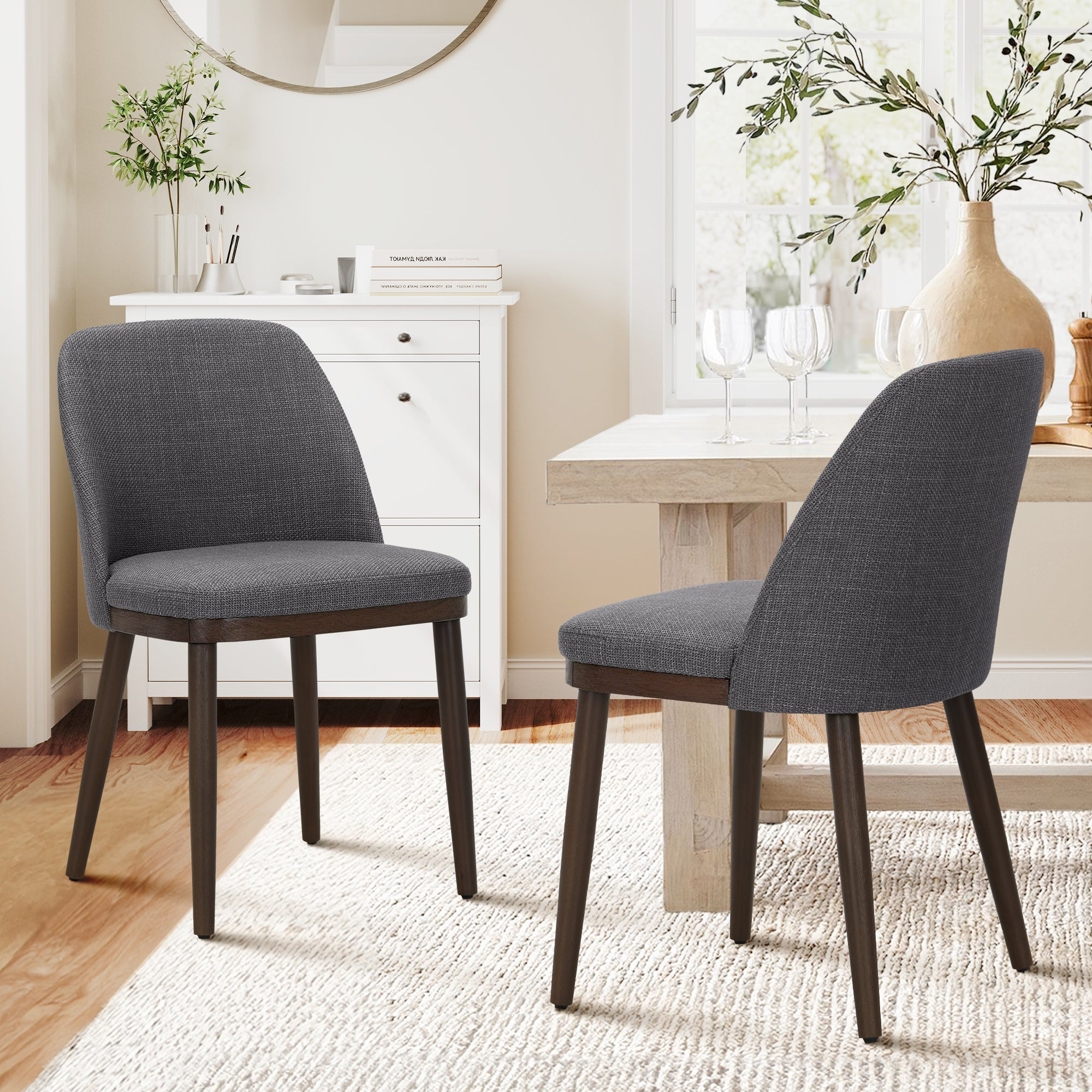 Due sedie imbottite grigio scuro con gambe in legno scuro si trovano in una sala da pranzo luminosa su un tappeto bianco.