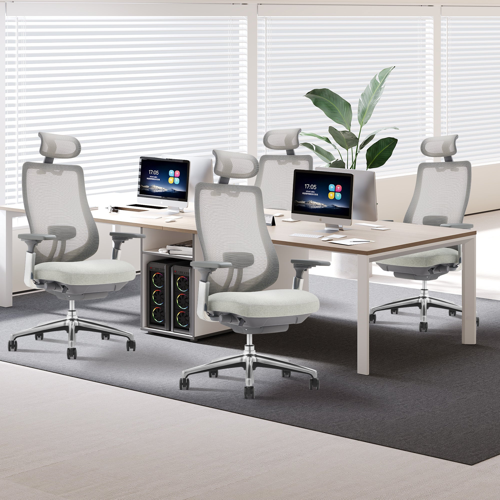 Modernes Büro mit Holztisch, ergonomischen grauen Stühlen, Computern und einer großen Grünpflanze vor Jalousien.