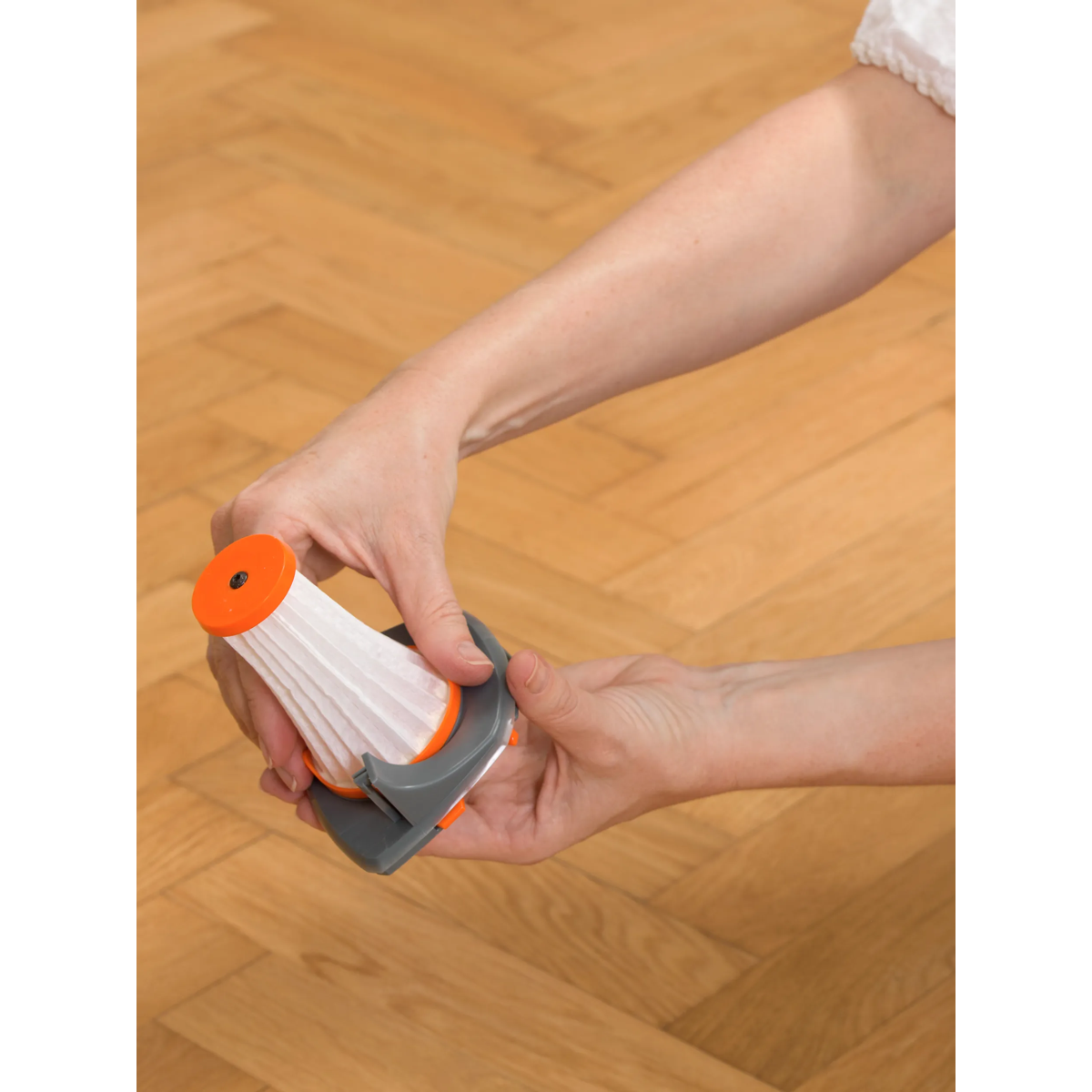 Manos sostienen un pequeño filtro de aspiradora con plástico naranja y gris sobre un piso de madera.