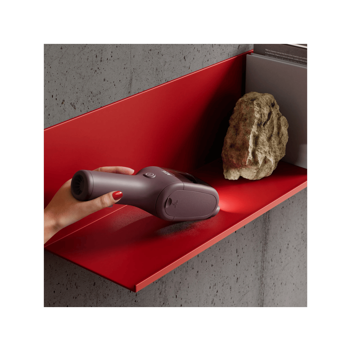Una mano sostiene una aspiradora de mano en una estantería roja contra una pared de hormigón gris.