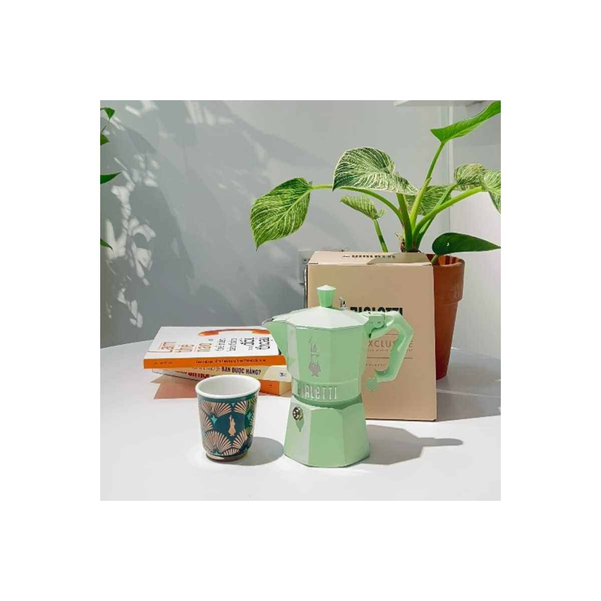 Una moka verde, una tazza decorata, libri e una pianta in vaso su un tavolo bianco contro una parete.