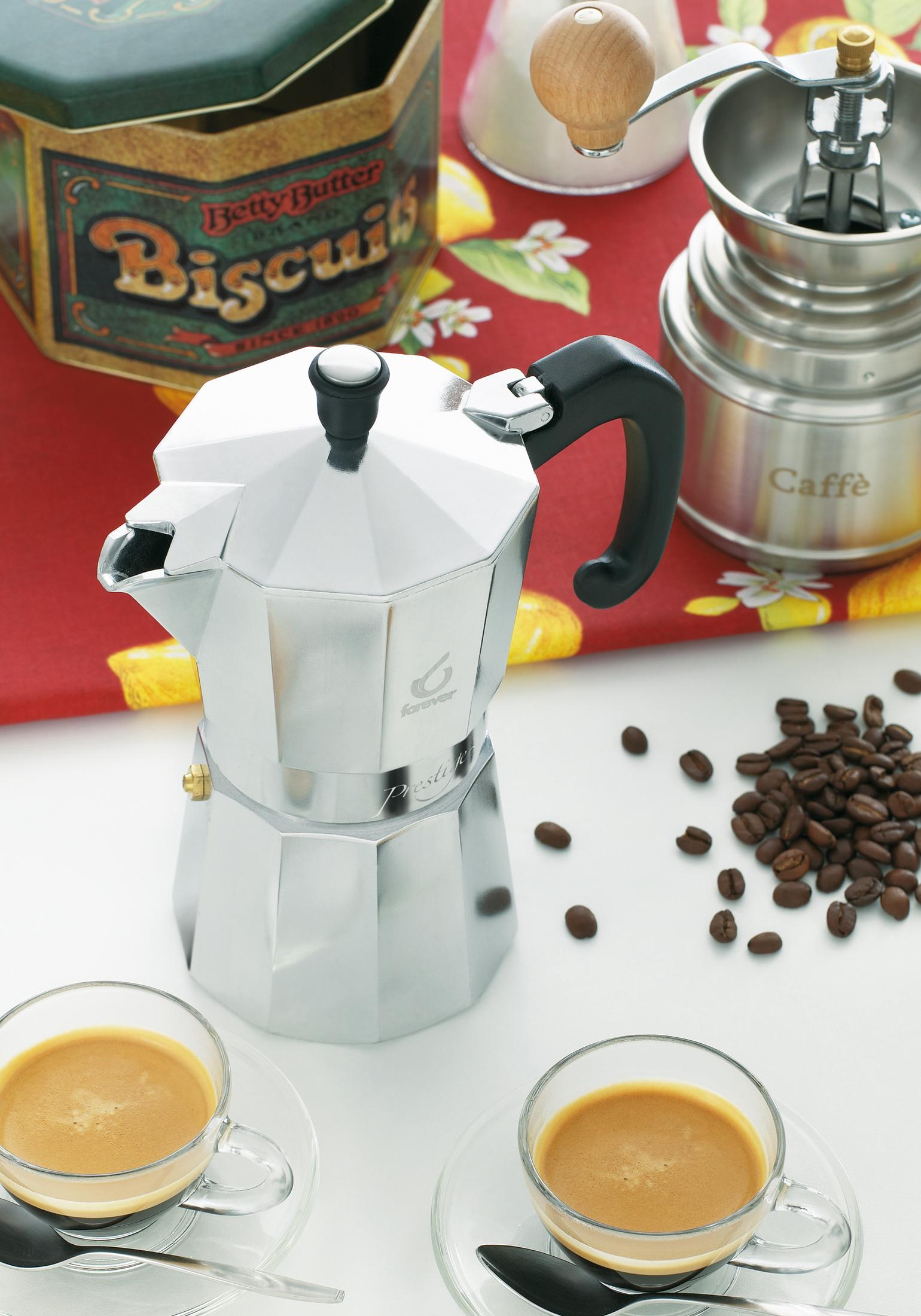 Una moka argentata, chicchi di caffè, due tazzine di espresso, una scatola di caffè e un macinino su un panno.