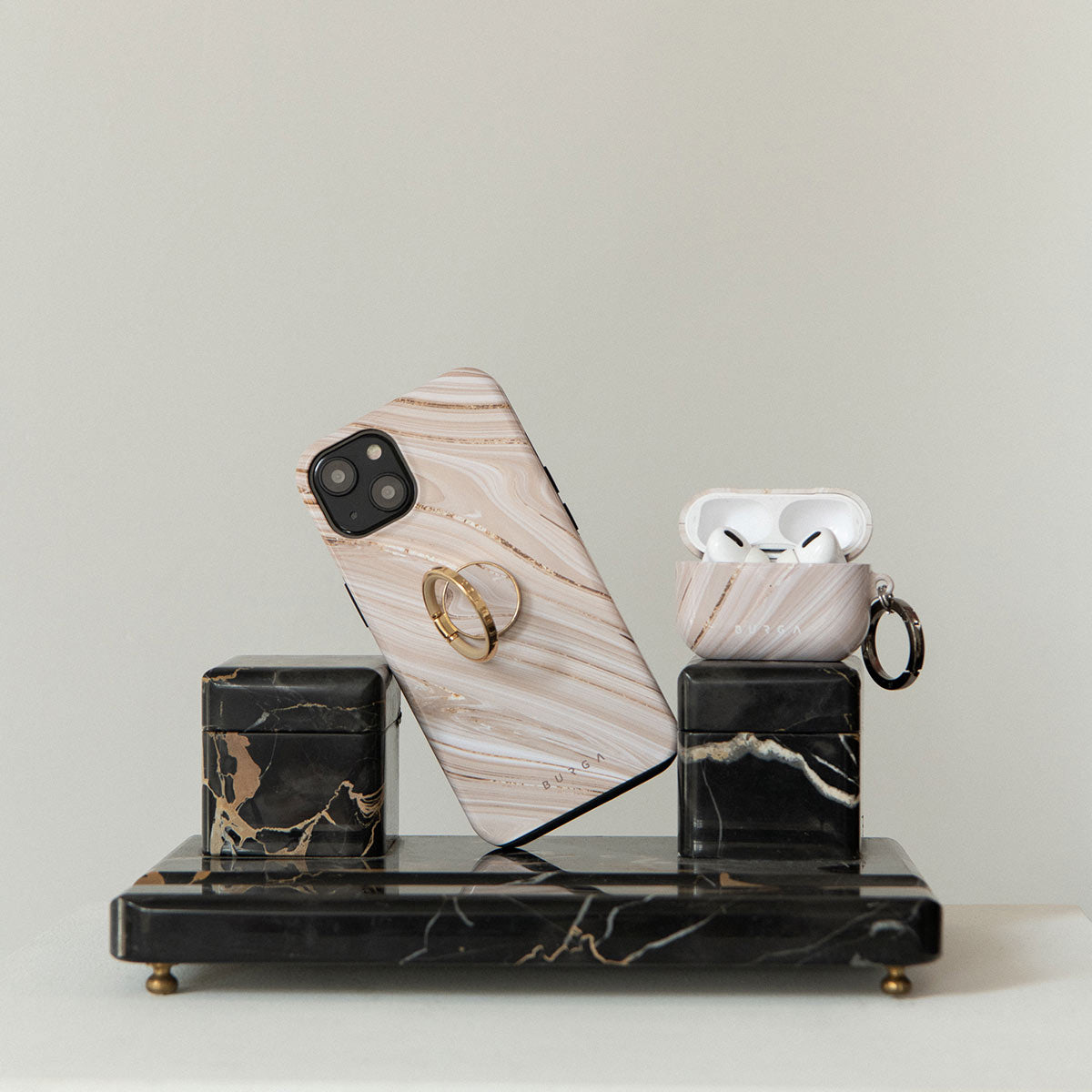 Handyhülle und AirPods-Case in beigem Marmordesign auf zwei schwarzen Marmorblöcken und einem Tablett.