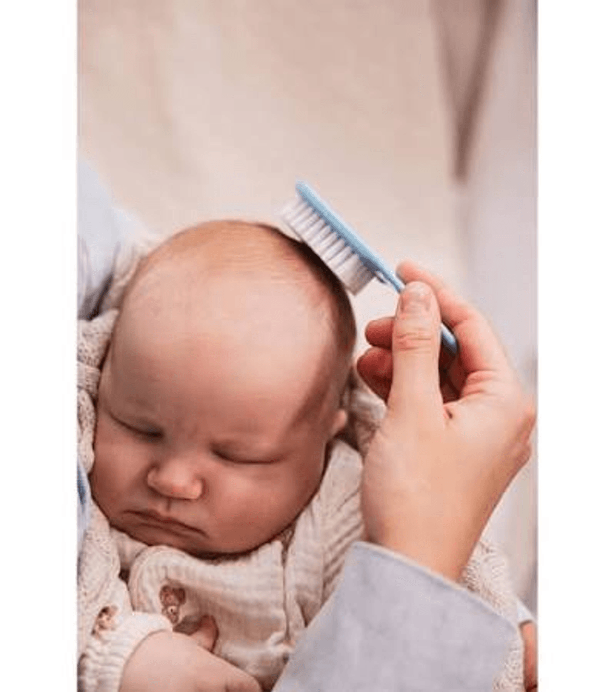 Ręka delikatnie czesze główkę śpiącego dziecka małą niebieską szczotką.