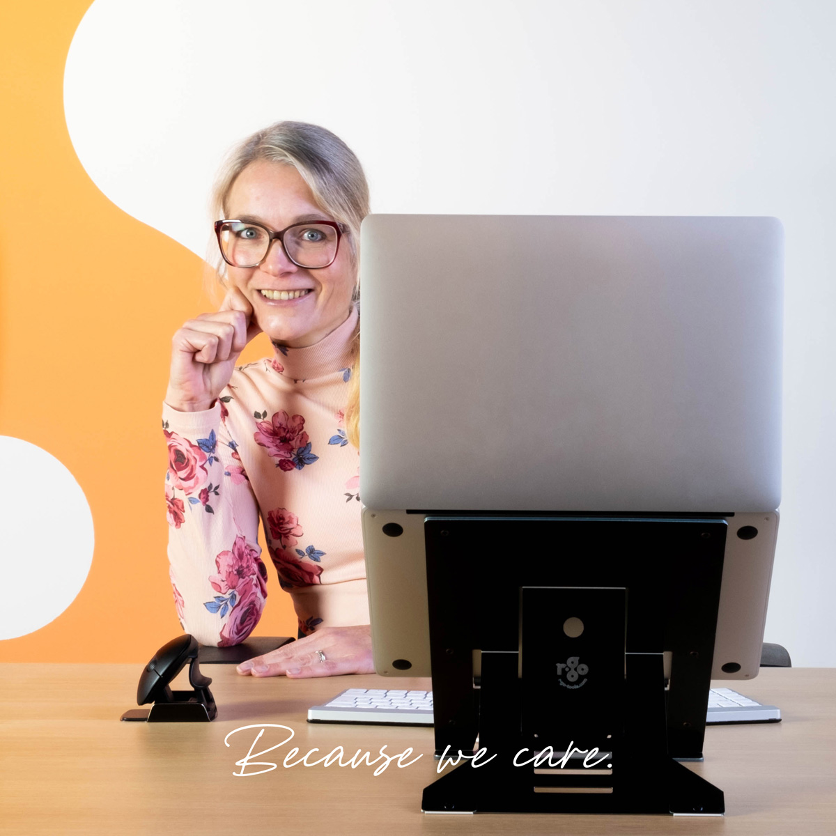 Eine blondhaarige Frau mit Brille und Blumenpulli sitzt am Schreibtisch hinter einem Laptop und lächelt.