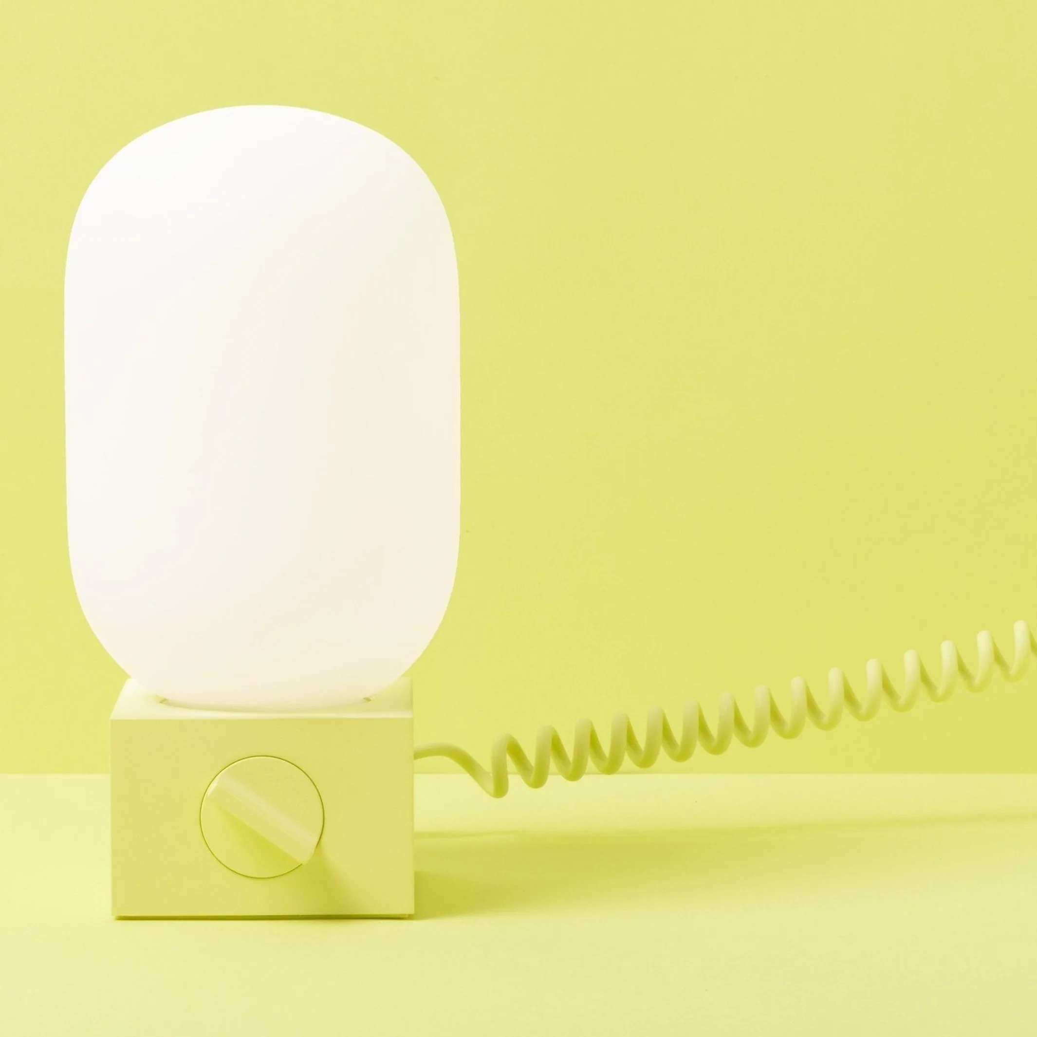 Biała lampa w kształcie kapsułki z żółtą podstawą i zwiniętym przewodem na żółtym tle.