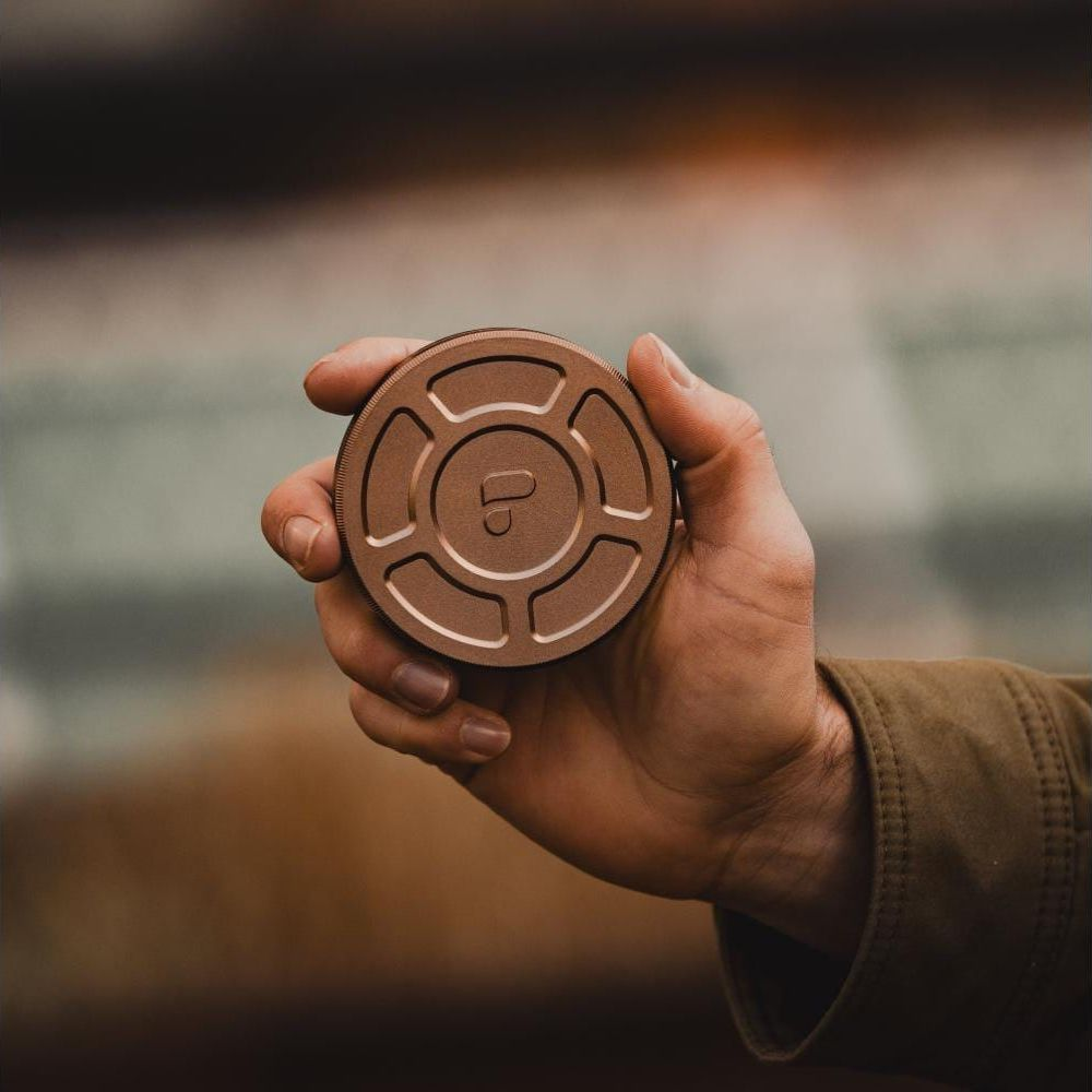 Eine Hand hält eine bronzefarbene, runde Dose mit einem Logo im Fokus, der Hintergrund ist verschwommen.