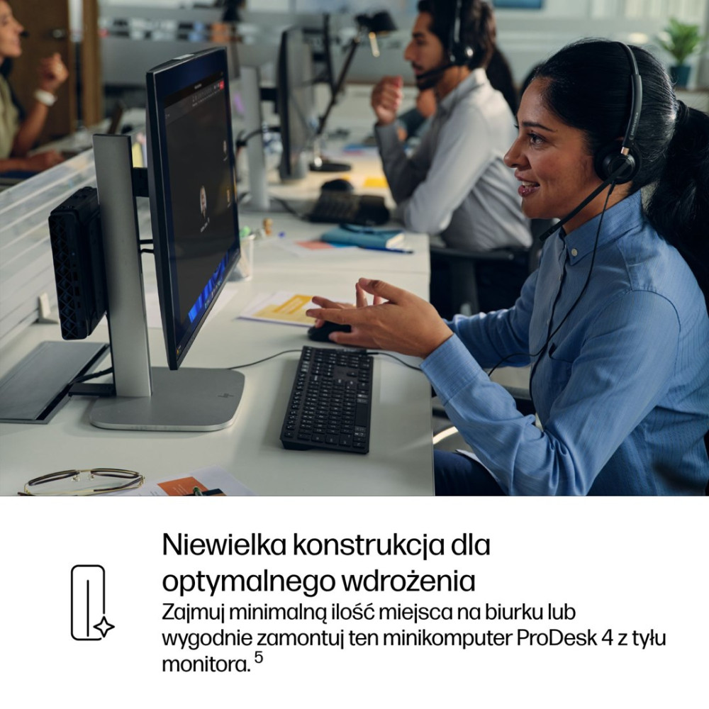 Kobieta w słuchawkach pracuje przy biurku z komputerem, a w tle widać innych pracowników biurowych.