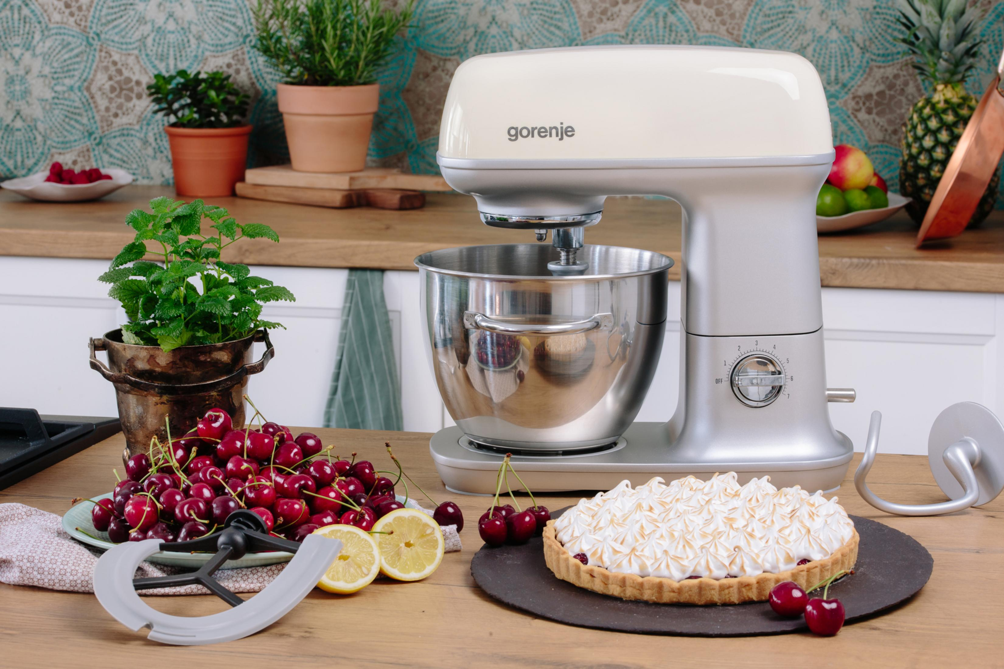 Eine cremefarbene Küchenmaschine steht auf einer Holzarbeitsplatte neben frischen Kirschen, Zitronen und einem Kuchen.