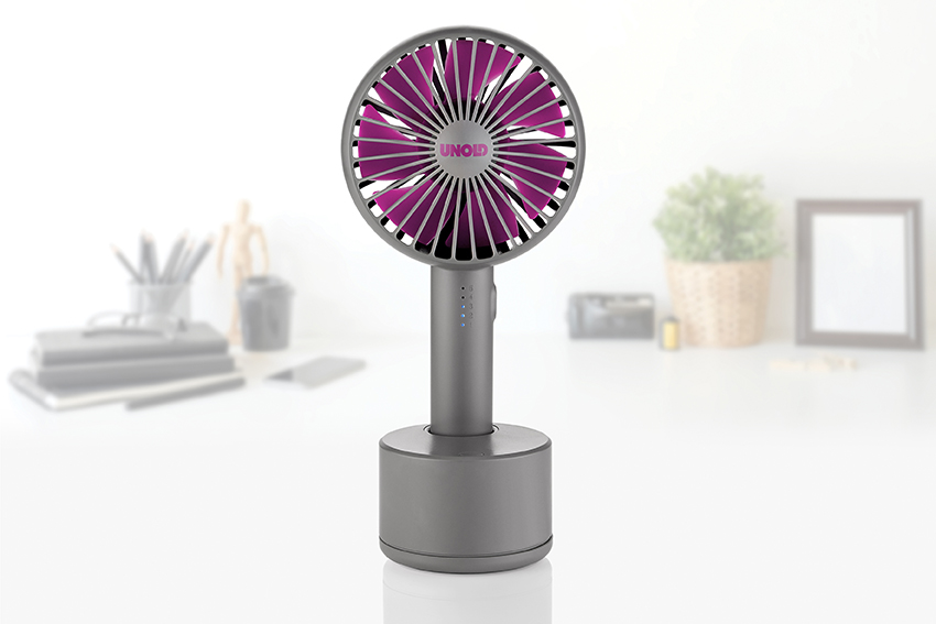 Grauer Handventilator mit pinken Rotorblättern auf einem Ständer vor verschwommenem Schreibtischhintergrund.
