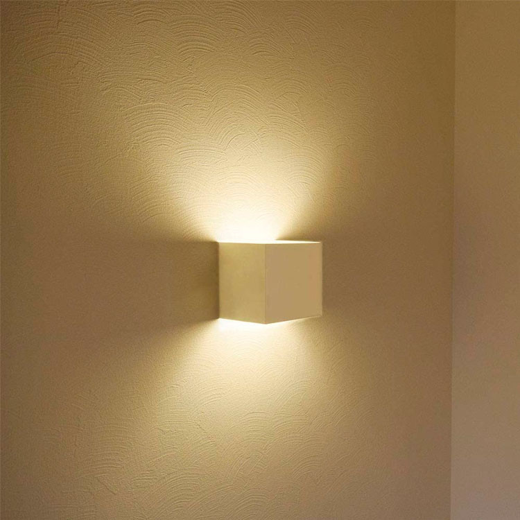 Una lámpara de pared blanca y cúbica moderna proyecta luz cálida en una pared texturizada.