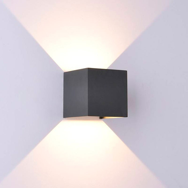 Una lámpara de pared cúbica negra proyecta haces de luz triangulares brillantes hacia arriba y hacia abajo en una pared blanca.