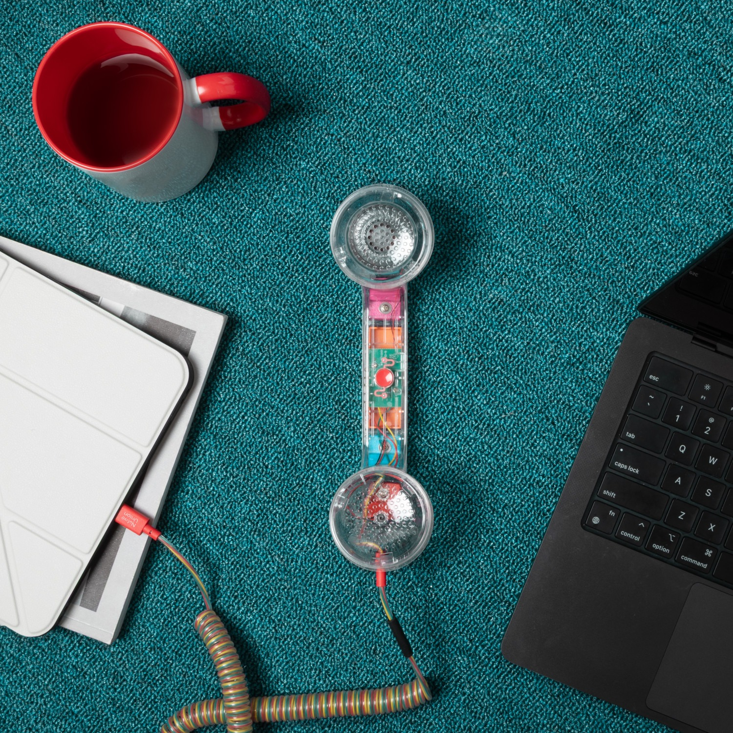 Na niebieskim kocu leży czerwony kubek, tablet, przezroczysta słuchawka telefoniczna i laptop.