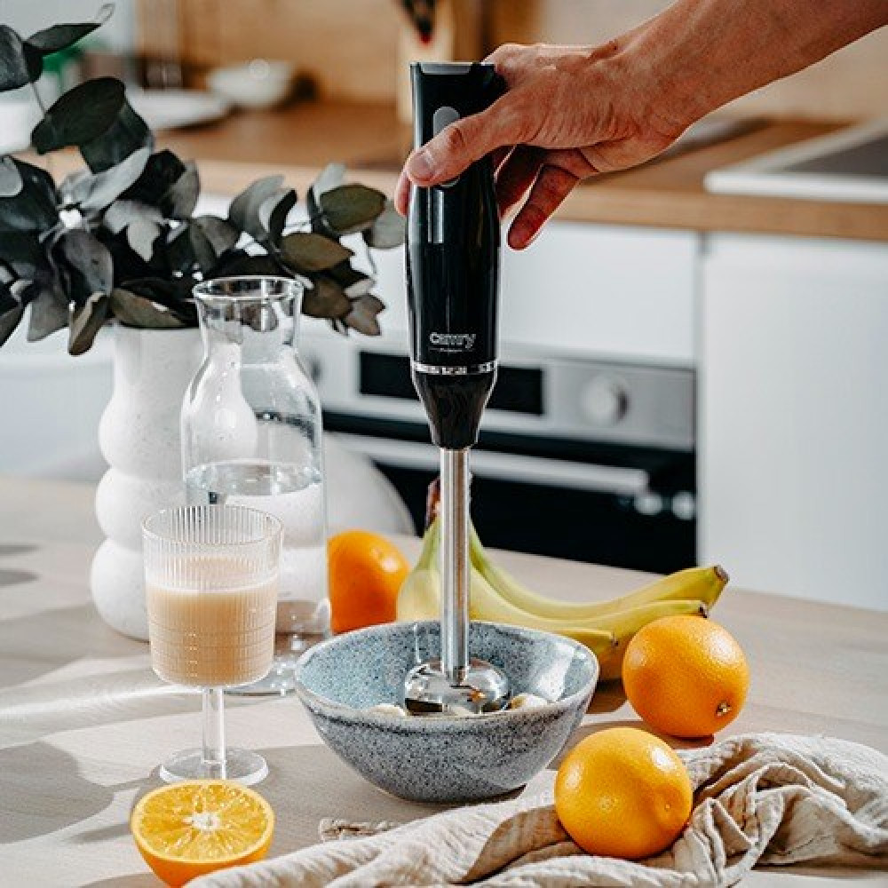 Ręka trzyma czarny blender ręczny nad miską z owocami na blacie kuchennym z pomarańczami, bananami i smoothie.
