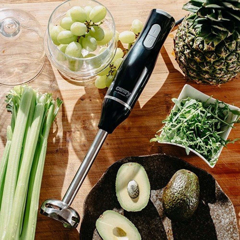 Czarny blender ręczny na drewnianym stole z winogronami, ananasem, selerem, rukolą i awokado.