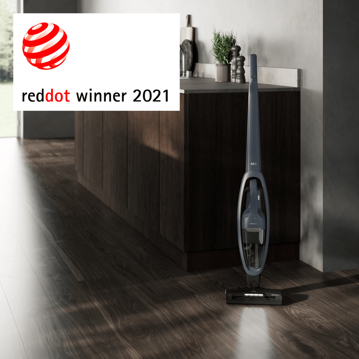Ein grauer AEG-Staubsauger steht auf einem dunklen Holzboden in einer modernen Küche mit Red Dot Award Siegel.