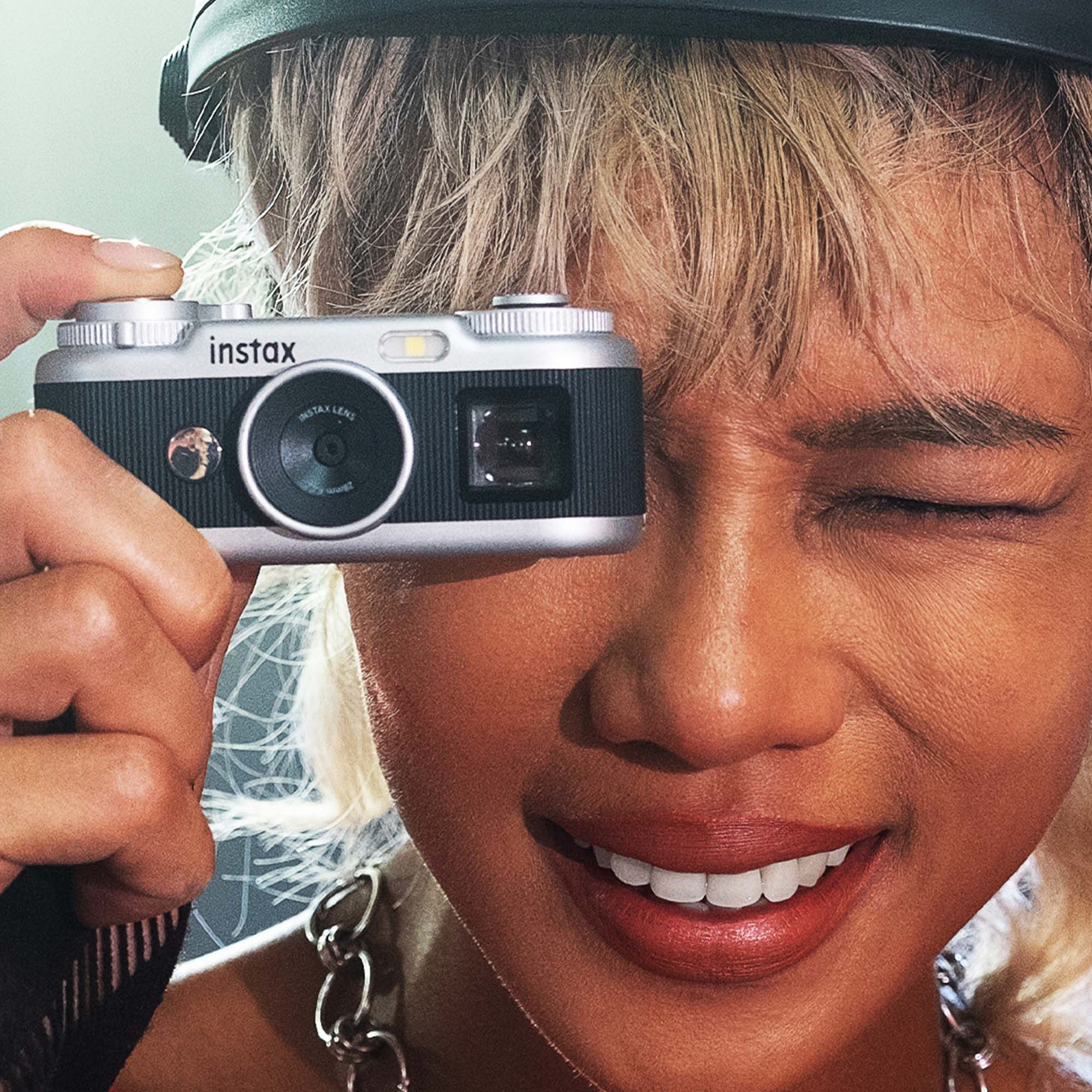 Sarışın, güler yüzlü bir kişi gözünün önüne gümüş renkli bir Instax kamera tutuyor.