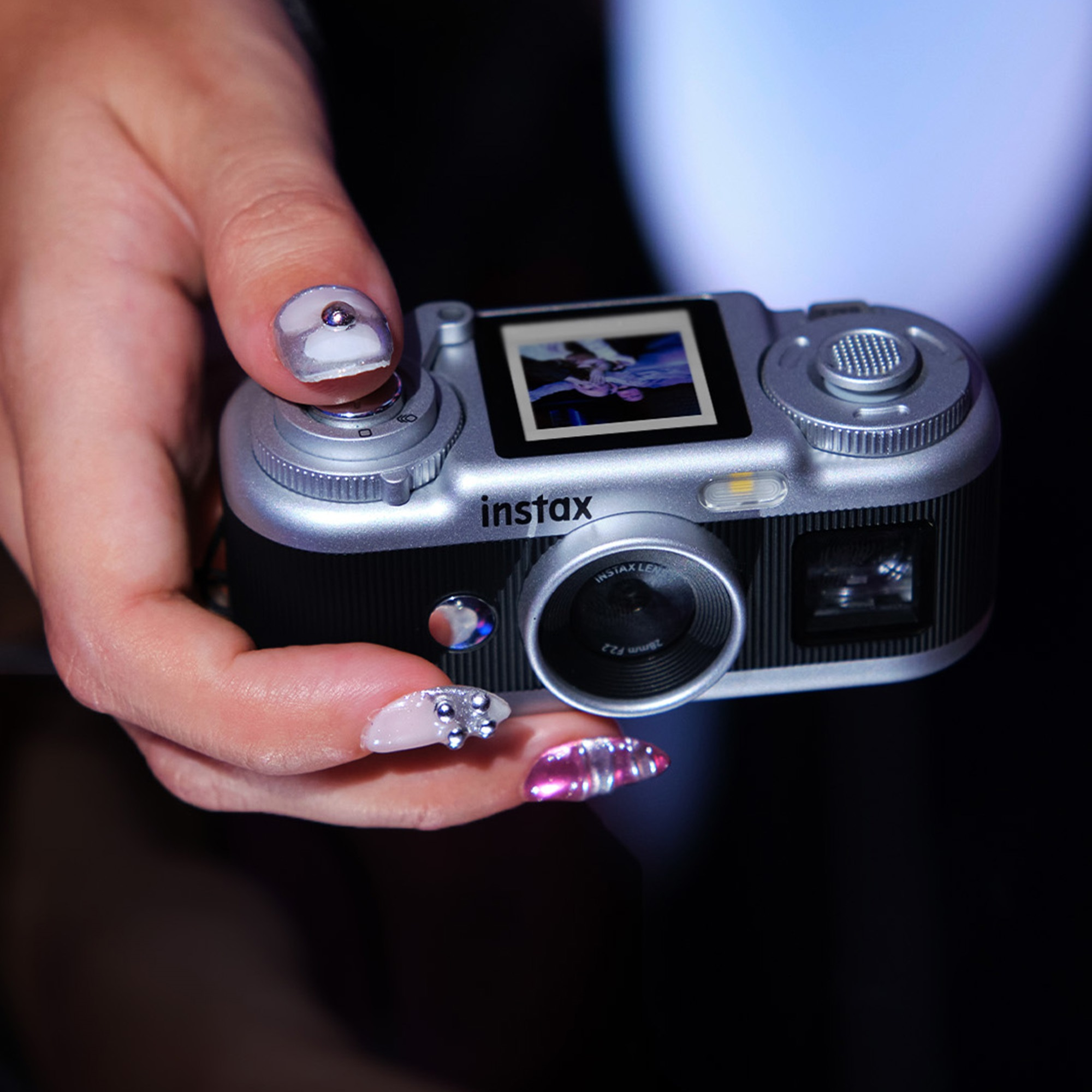 Bir el, küçük ekranlı ve süslü tırnaklı gümüş ve siyah bir Instax kamera tutuyor.