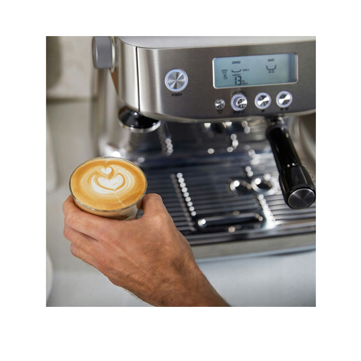 Eine Hand hält einen Latte mit Herzmuster vor einer Edelstahl-Espressomaschine.