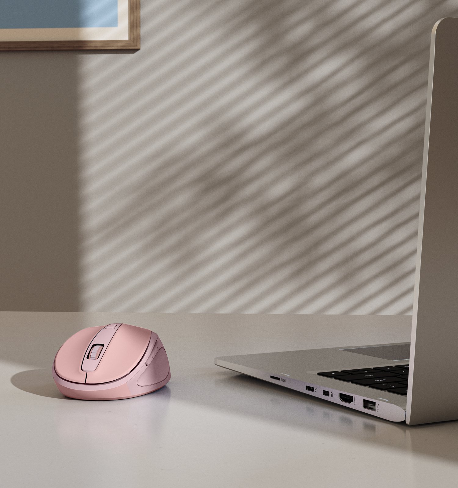Eine rosa Funkmaus liegt neben einem silbernen Laptop auf einem Schreibtisch mit Jalousienlicht.