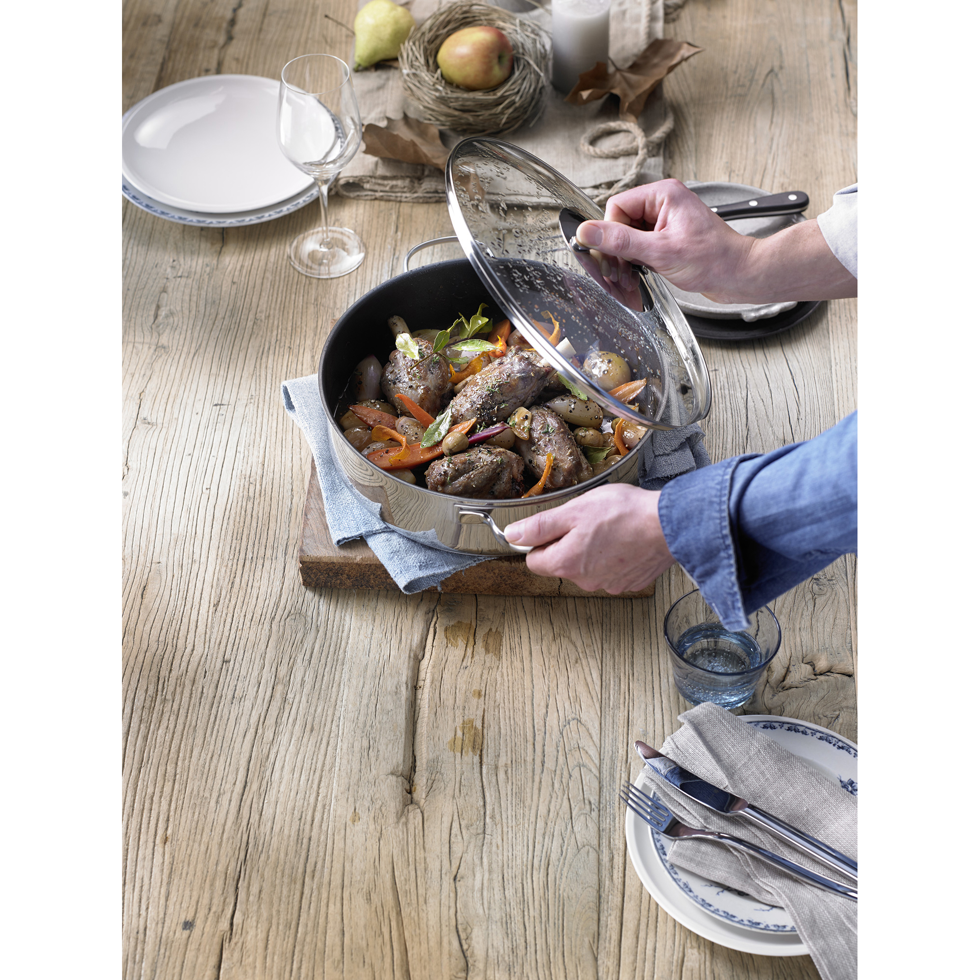 Person öffnet den Deckel eines Topfes mit Fleisch und Gemüse auf einem Holztisch.