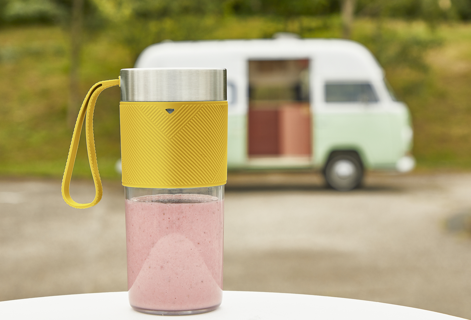 Ein gelb-silberner Becher mit rosafarbenem Smoothie steht auf einem Tisch vor einem retro Campervan.