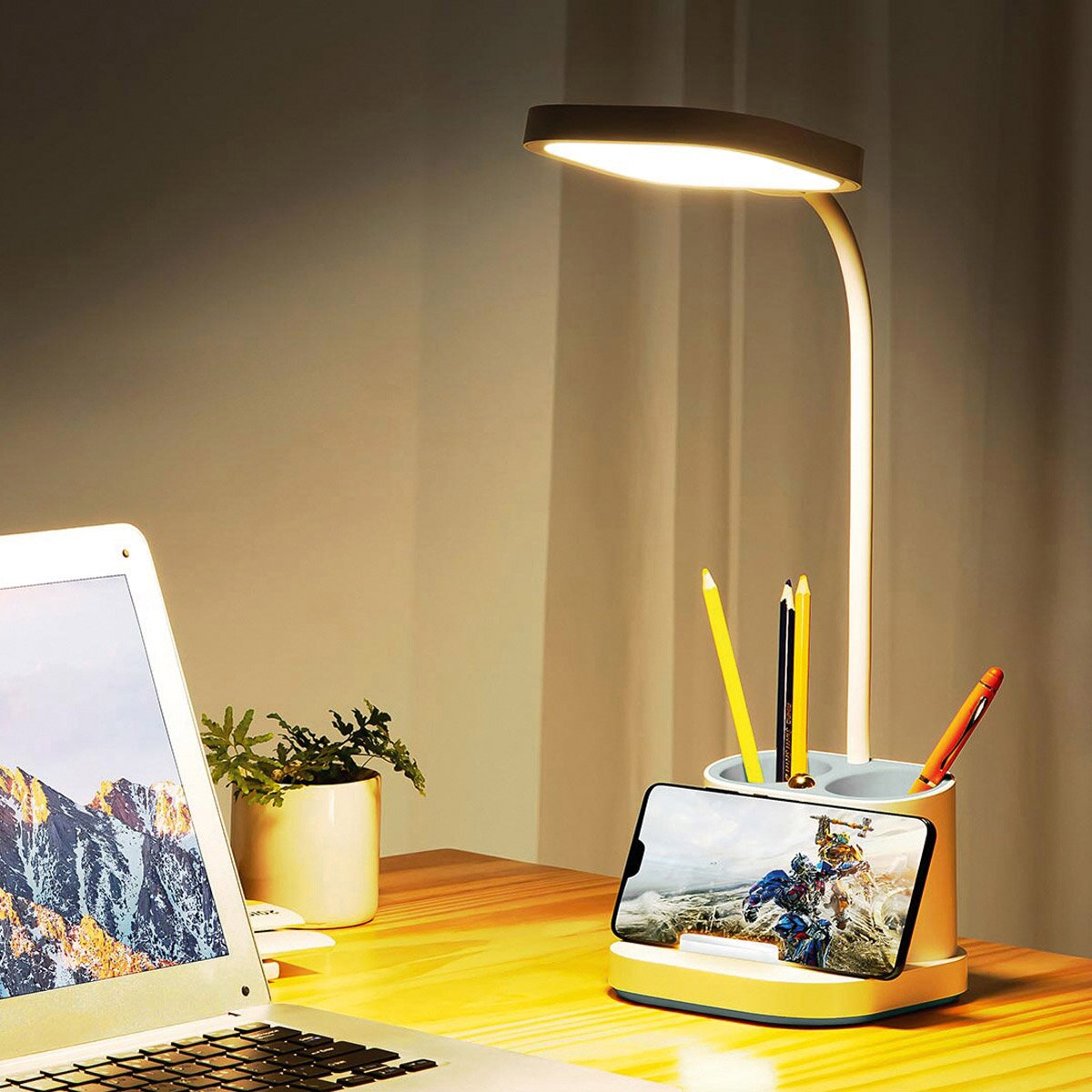 Eine Schreibtischlampe leuchtet auf einem Holztisch mit Laptop, Stiftehalter, Smartphone und einer kleinen Topfpflanze.