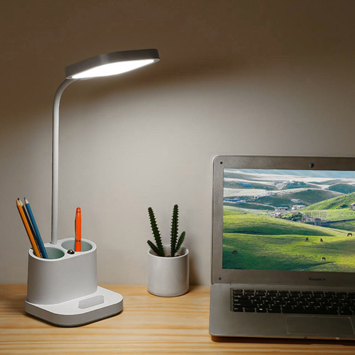 Eine weiße Schreibtischlampe mit Stiftehalter leuchtet neben einem Laptop und einer kleinen Pflanze.